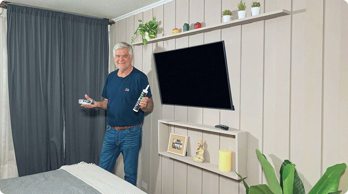 un hombre sostiene 2 tipos de adhesivos en las manos y está parado frente a un muro que acaba de renovar con listones pegados de MDF y unas repisas para decoración.