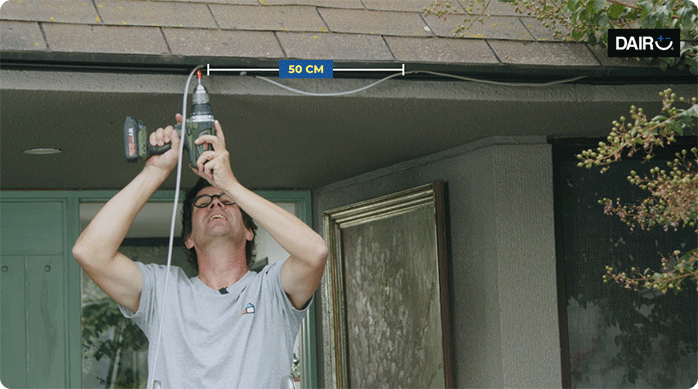 un hombre usa un atornillador eléctrico inalámbrico para poner las grampas para sostener la tira de luces led en el alero de la casa.