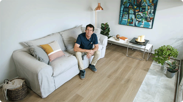 un hombre sentado en un sofá, en un living en el que se acaba de instalar piso vinílico con textura madera y piedra, combinando sus texturas.