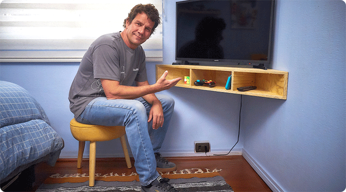 un hombre sentado en un piso muestra sonriente el nuevo mueble esquinero para la tv que acaba de instalar en un dormitorio.