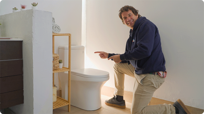 un hombre con polerón azul y pantalón beige hincado en un baño señala el WC de 1 pieza que acaba de instalar.