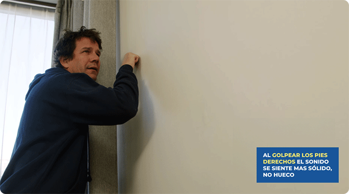 un hombre con polerón azul golpea un muro de yeso cartón para identificar dónde están los pies derechos.