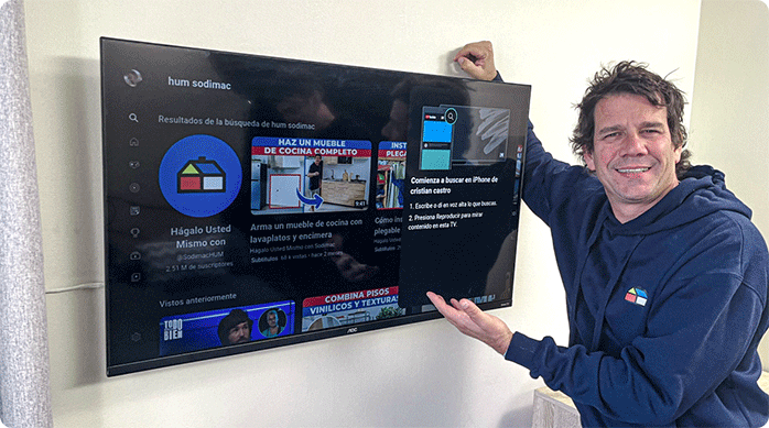 un hombre con polerón azul muestra el televisor que acaba de colgar a un muro de yeso cartón con un soporte para tv.