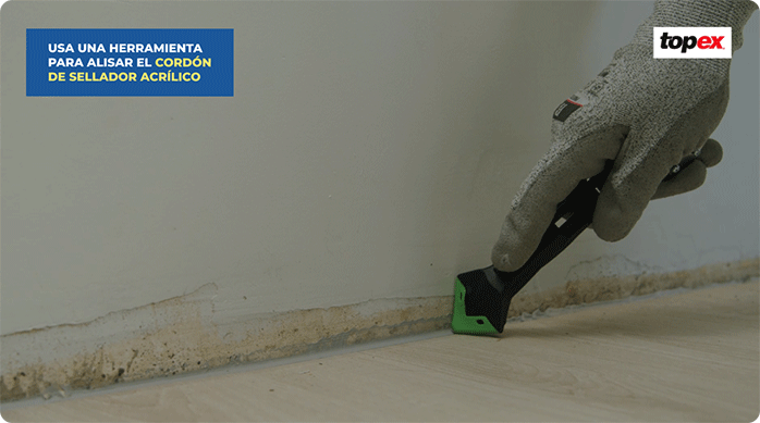 una persona pasa una herramienta para alisar el cordón de sellador acrílico en el borde del piso vinílico adhesivo.
