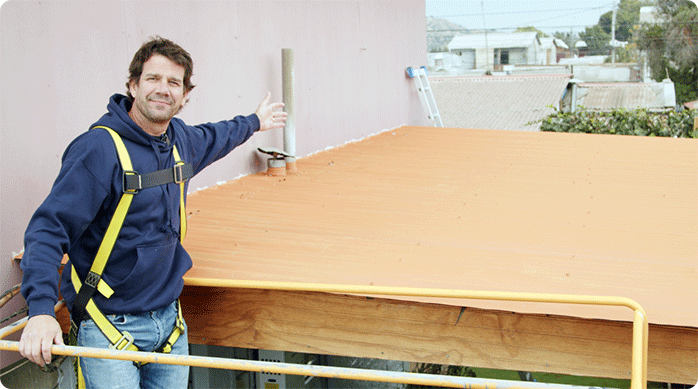 un hombre con arnés y cabo de vida subido a un andamio muestra orgulloso el techo de zinc que acaba de pintar con pintura anticorrosiva.
