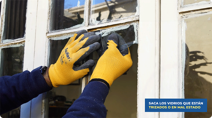 una persona con guantes de seguridad retira los vidrios rotos de una ventana de madera.