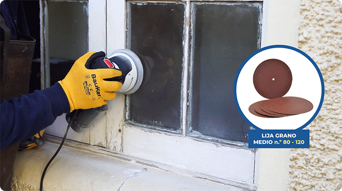 un hombre con guantes de seguridad pasa una lijadora roto orbital con disco grano medio en la madera de la ventana de madera para eliminar pintura antigua.
