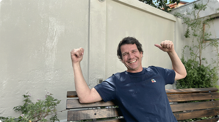un hombre sonriendo sentado en una banca e indicando un muro exterior que acaba de renovar con pintura látex.