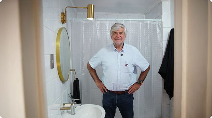 un hombre mayor con las manos en la cintura sonriendo, parado en un baño pequeño que acaba de renovar sin obra.