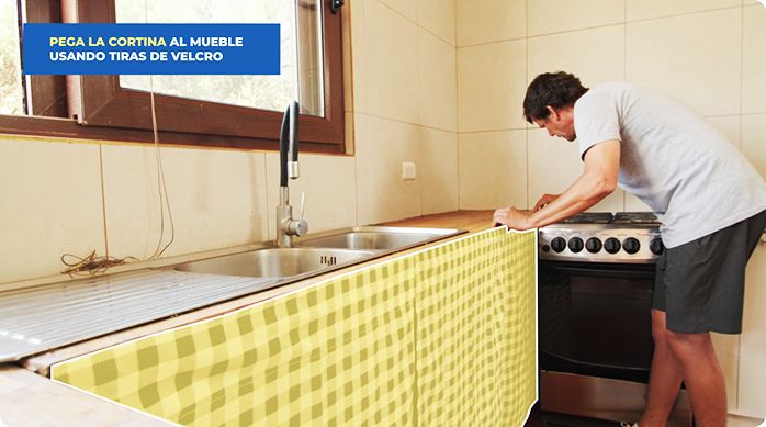 un hombre instala una cortina de cuadros para cubrir un mueble de cocina que no tiene puertas.